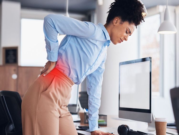 Photographie d'une femme debout devant son bureau, se tenant le bas du dos, penchée en avant, à cause d'une douleur aux lombaires.