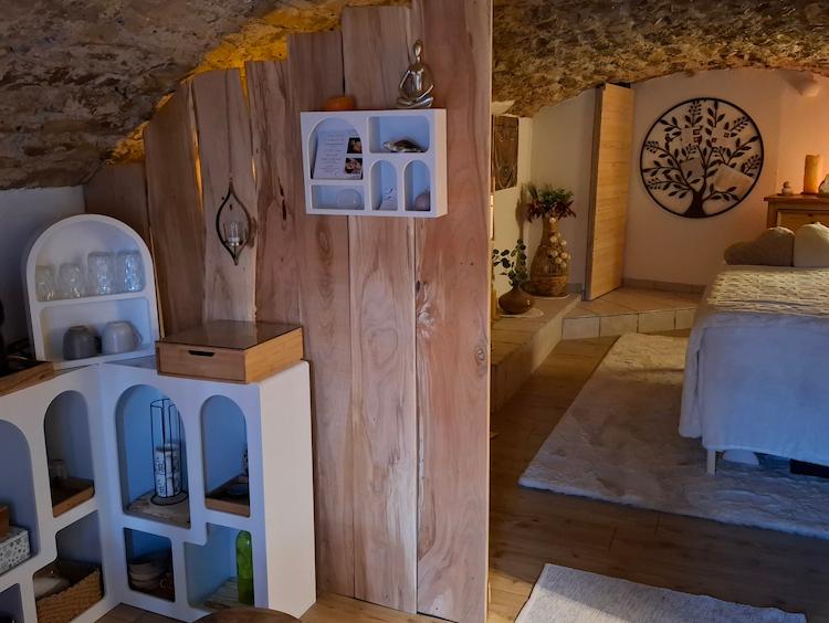 Photographie du salon depuis l'entrée. L'étagère de l'entrée est visible, et derrière un panneau en bois on peut appercevoir le lit de massage ainsi que les différentes décorations.