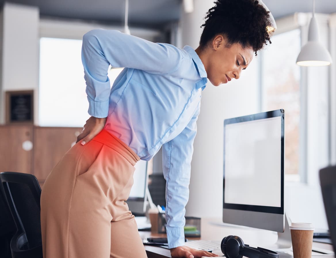 Photographie d'une femme debout devant son bureau, se tenant le bas du dos, penchée en avant, à cause d'une douleur aux lombaires.